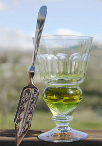 A reservoir glass filled with a naturally coloured verte absinthe, next to an absinthe spoon.