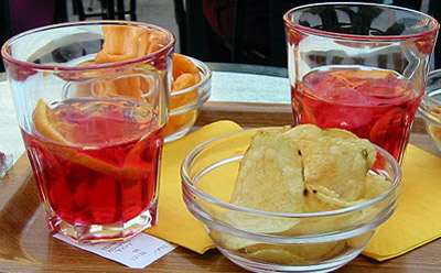 Two glasses of apéritifs (Campari with carbonated water) served with potato crisps and peanuts as appetizers.